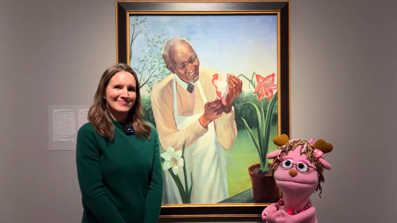 Two figures stand on each side of a portrait hung on a wall. The figure to the left wears a green sweater. The figure to the right is a pink jackalope puppet. The portrait is of G. W. Carver holding a lily flower.