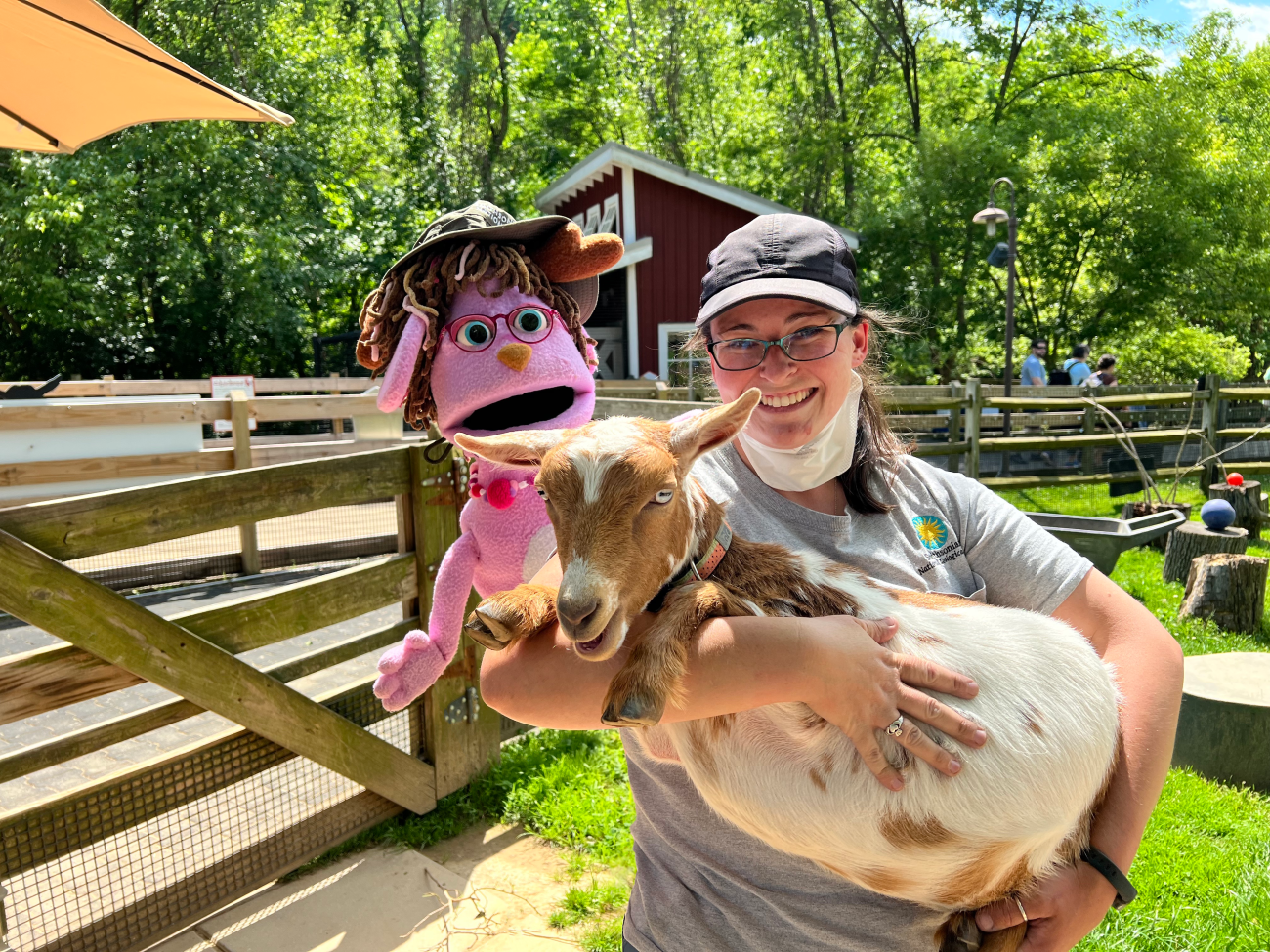 Zoo keeper Nikki stands while she holds brown and cream spotted Nigerian dwarf goat. Behind her left should a pink jackalope puppet smiles.