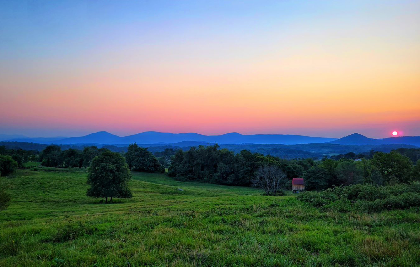 A sunrise over a rural, grassy area with trees and a low mountain range in the background