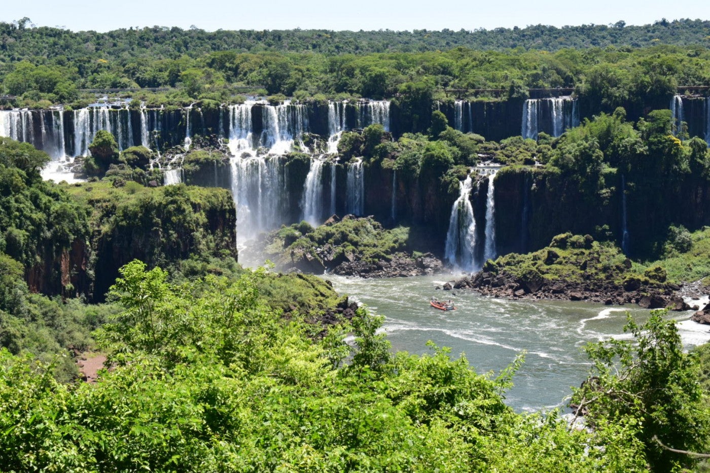 a large waterfall is seen from a distance. lush vegetation surrounds the area.