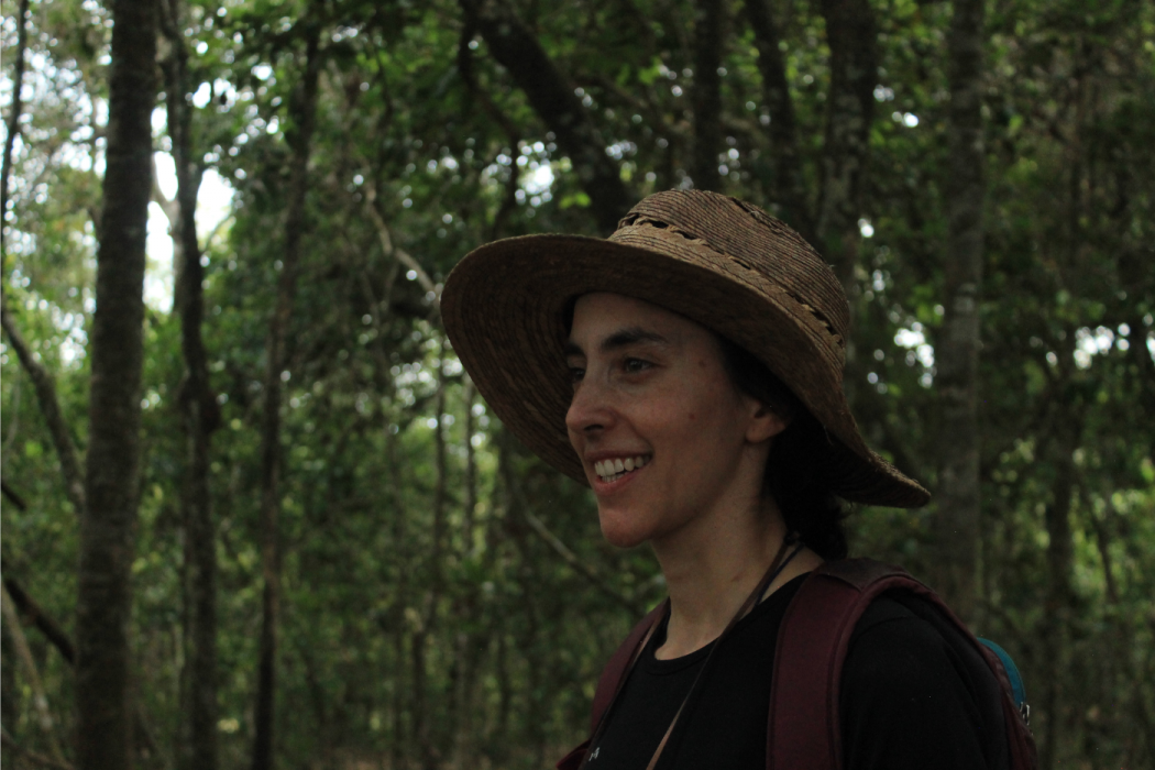 Photo of Juliana Velez Gomez in a forest. Juliana is a Caucasian Latina woman, with dark hair. She is wearing a broad brimmed hat and smiling.