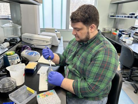 A photo of Sean Lyons in a lab setting. He is using a pipette to put fluid into a small vial. 