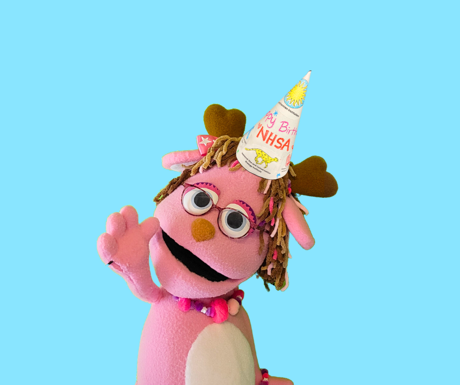 A pink jackalope puppet with her arm raised waving. She wears pink glasses and a birthday hat on the top of her head.