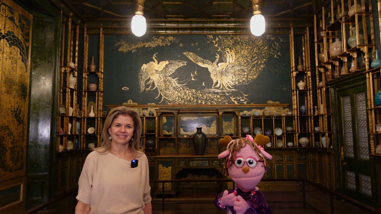 A woman stands next to a pink jackalope puppet in front of a wall with two painted gold peacocks. The room they are standing in is called the Peacock Room found at the National Museum of Asian Art.