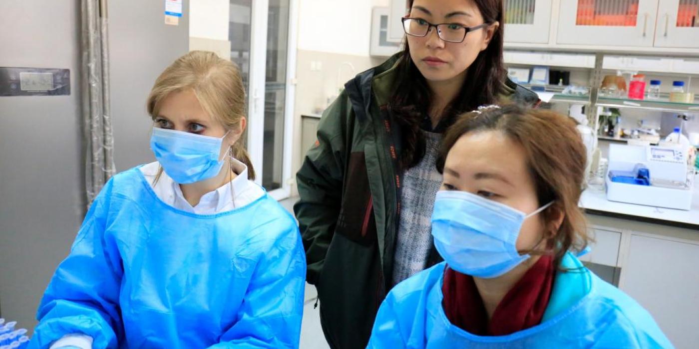 Three researchers, two wearing masks, gloves and scrubs, examine data on a computer