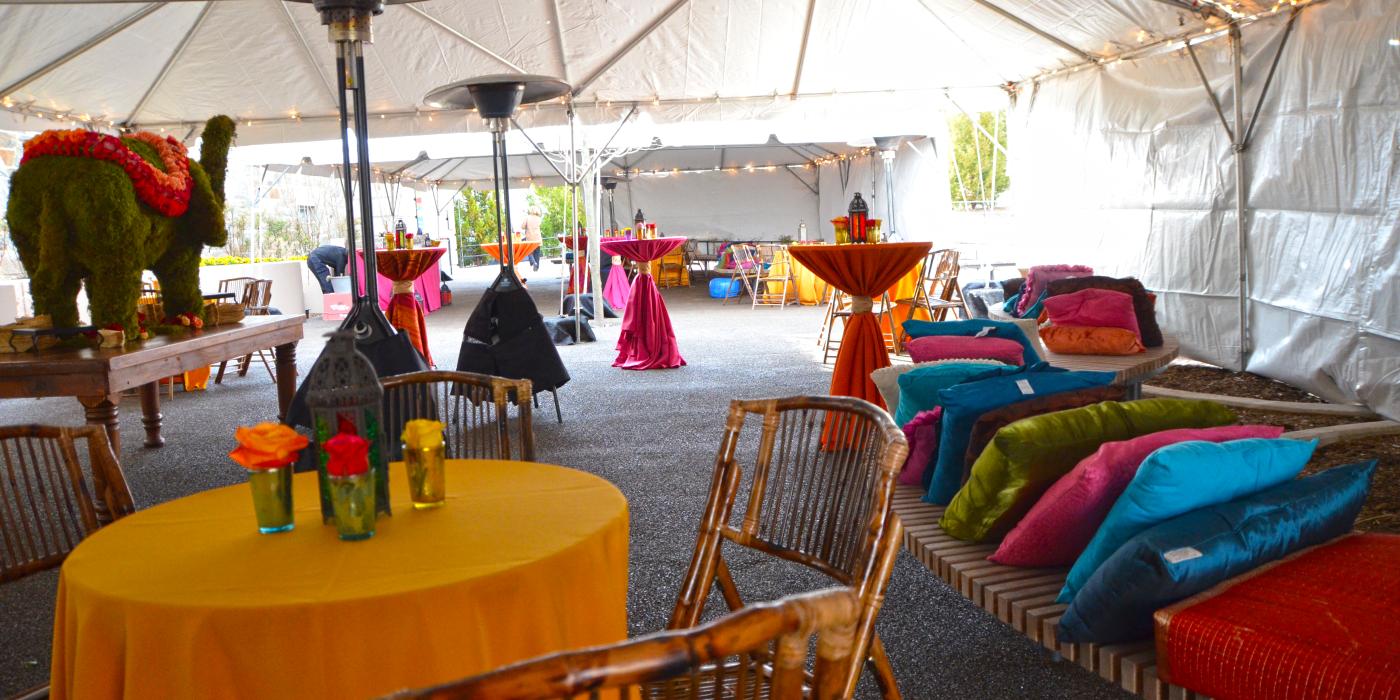 A colorful catered event set up under a large tent with cocktail tables, benches with pillows and lights